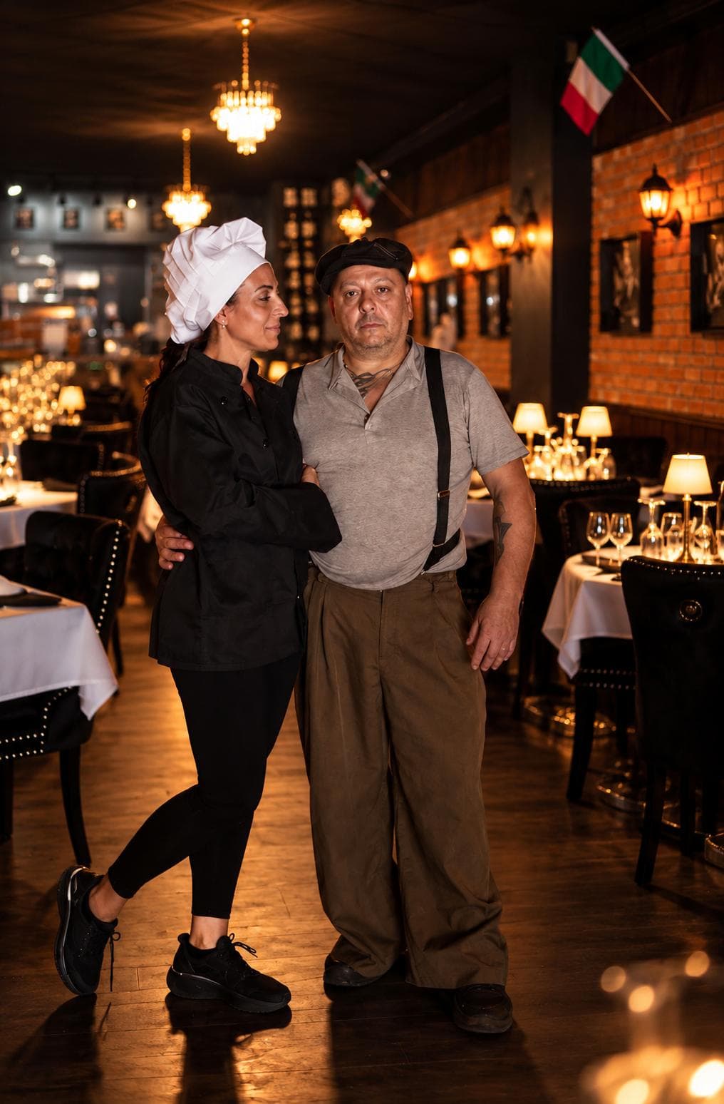 A chef in a black jacket and white toque beside a man in a flat cap and suspenders, in the dining room