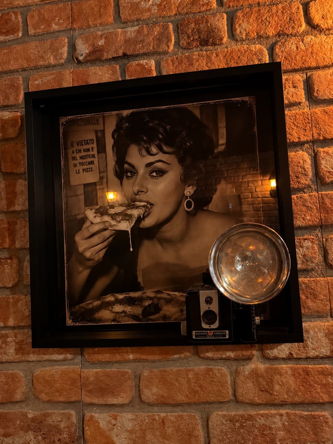 A framed sepia film still of a screen star biting into a slice of pizza, a vintage Brownie Hawkeye flash camera mounted on the frame, on the brick wall