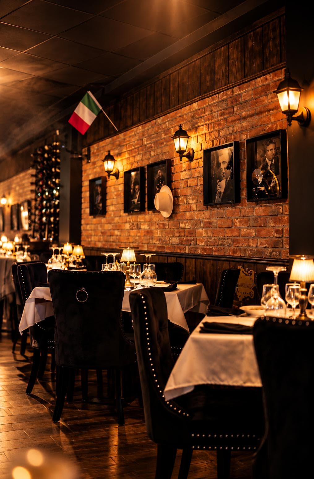 Lanterns, framed portraits and a white fedora on the brick wall, above tables with lamps and wine glasses