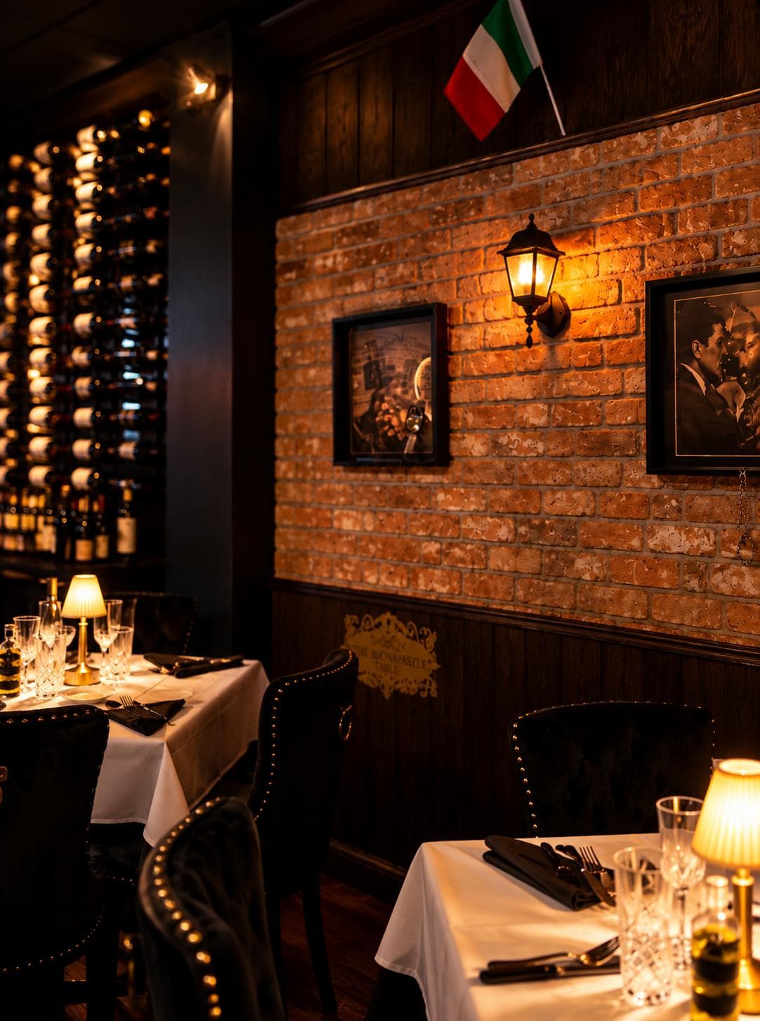 Tables with brass lamps by the lit wine wall, a lantern and framed portraits on the brick, an Italian flag above